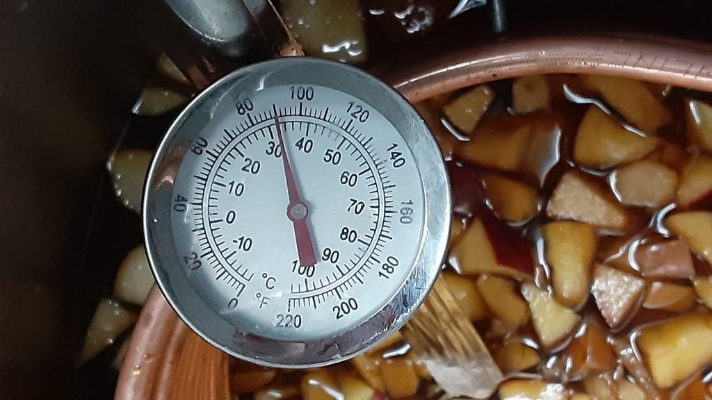 A picture of a thermometer showing 85 degrees F (31 C), in the background some brown liquid with cubes of apple and pear floating.