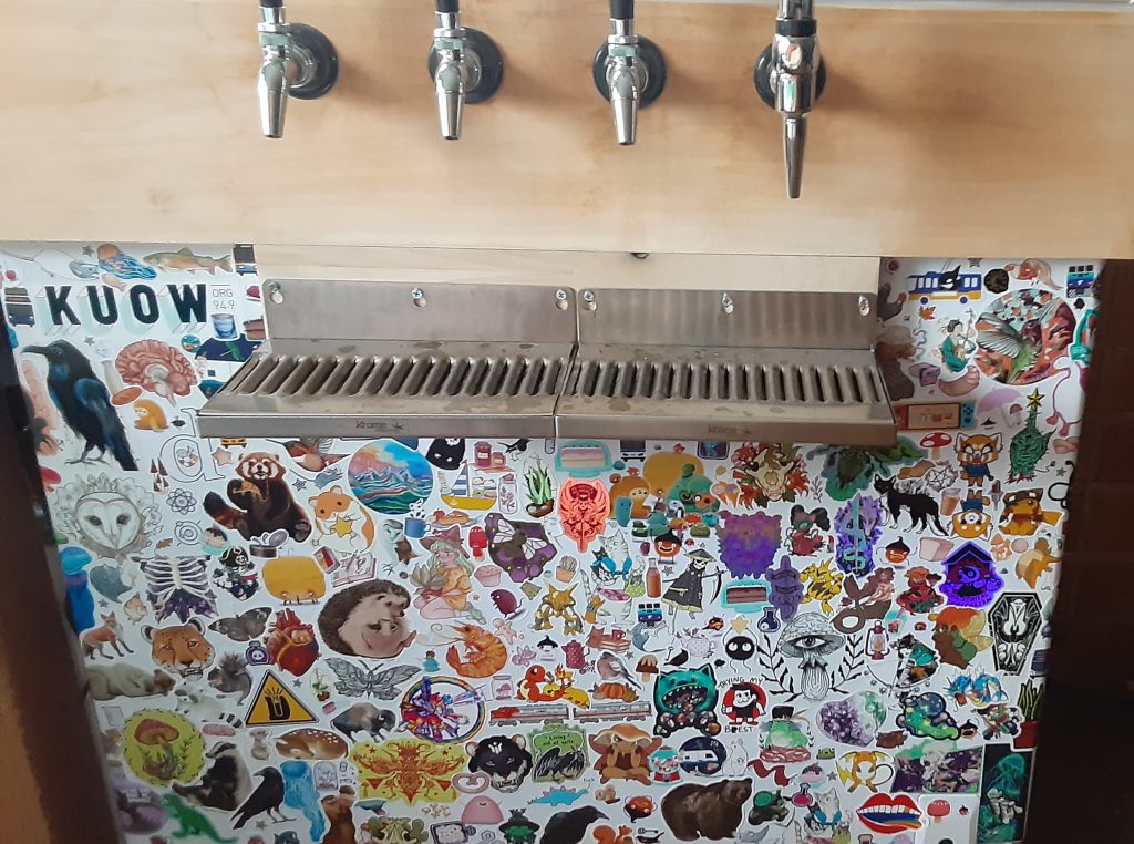 Photo of a kegerator with 4 taps, a drip tray, and a fridge part covered in stickers.